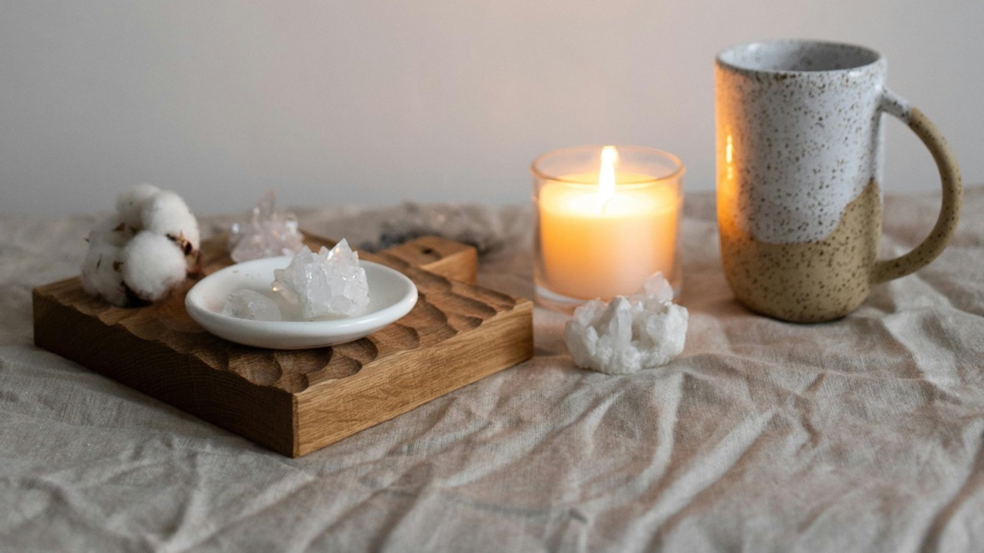 A lit candle, cotton, crystals, and ceramic mug on a linen cloth, creating a serene night skincare routine atmosphere.