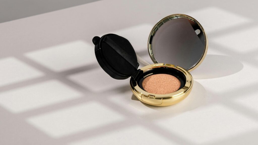 Editorial flatlay of gold compact makeup in soft neutral light, representing glow-focused makeup