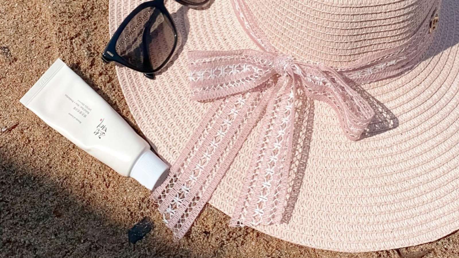 Photo for The Summer Makeup Edit of a sun hat, sunscreen tube, and sunglasses on sand.