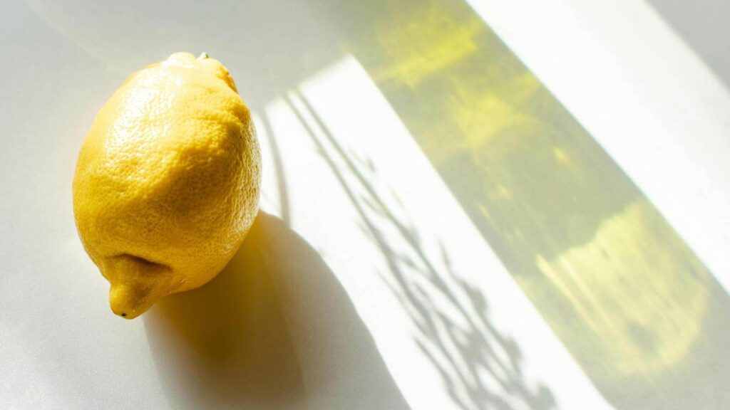Lemon on soft background with sun shadows representing why your vitamin c isn’t making your skin look brighter.