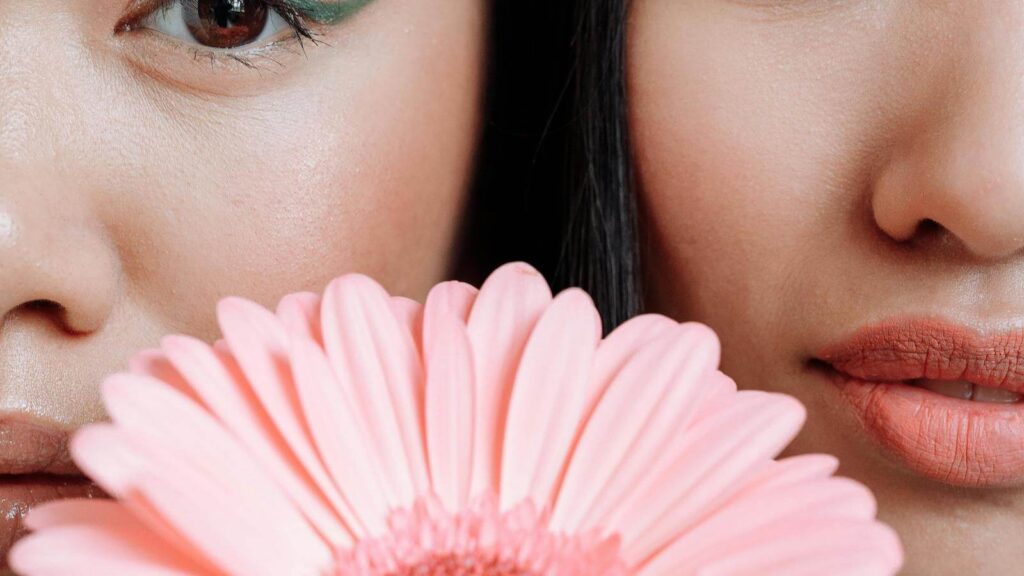 Close-up of two women's faces with dewy makeup, partially hidden behind a pink flower, illustrating best dewy primers for glowing skin