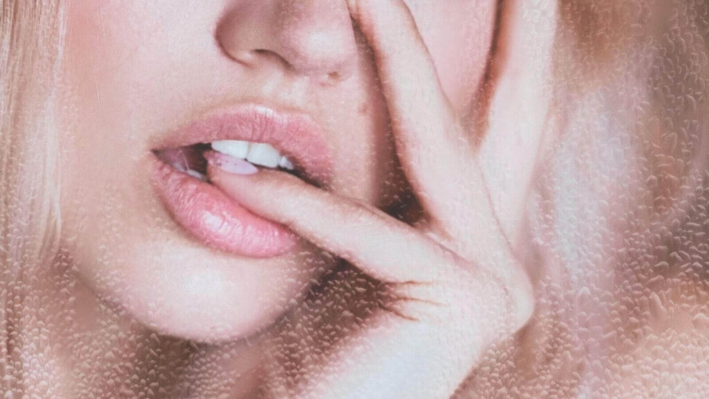 Close-up of woman gently biting finger, demonstrating soft glossy lips for a glowing makeup look in natural light.