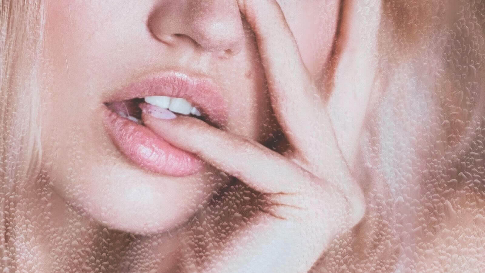 Close-up of woman gently biting finger, demonstrating soft glossy lips for a glowing makeup look in natural light.