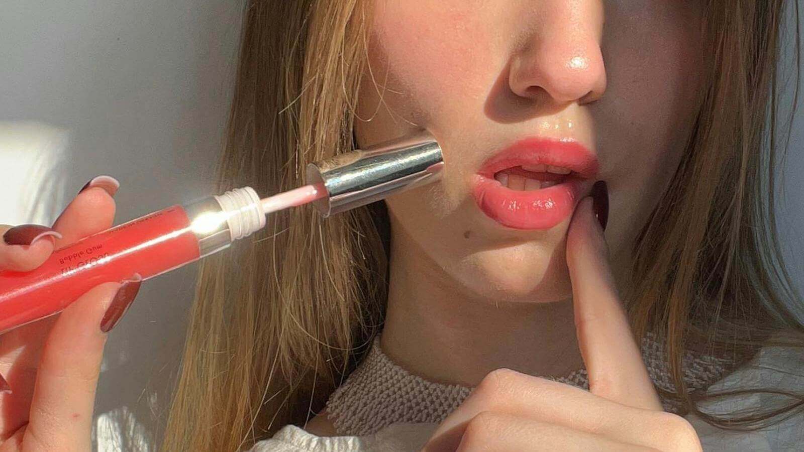Close-up of woman applying coral lip gloss, demonstrating soft glossy lips for a glowing makeup look in natural light.